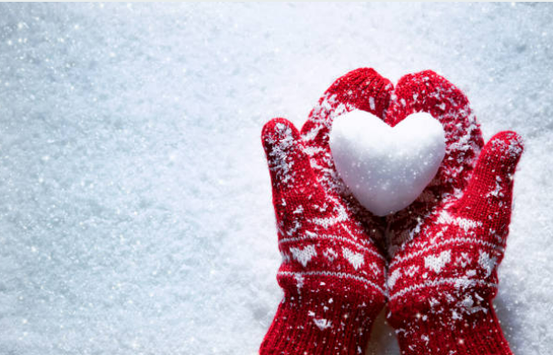 Two hands together wearing red and white mittens holding a heart made out of snow.