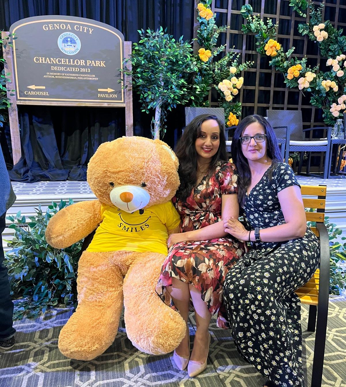 Two people sitting on a bench beside a giant teddy bear.