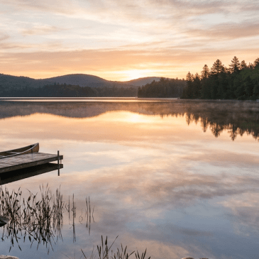 Sunrise over a still lake with a dock and canoe, surrounded by forest and hills.